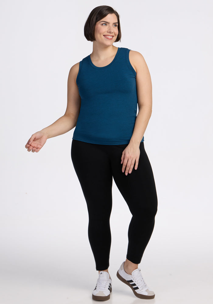 A woman with short brown hair smiles while wearing the Woolx Ava Tank Top in Majolica Blue, paired with black leggings and white sneakers with black accents, standing against a plain white background.