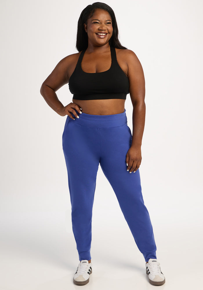 Woman smiles in Woolx Madden Joggers - Royal Blue and a black sports bra, paired with white sneakers on a white background.