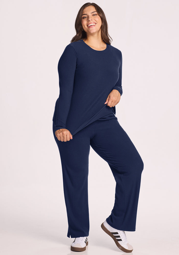 Smiling woman in Woolx Hadley Ribbed Crew - Ocean Cavern, pants, and white sneakers, holding her shirt.