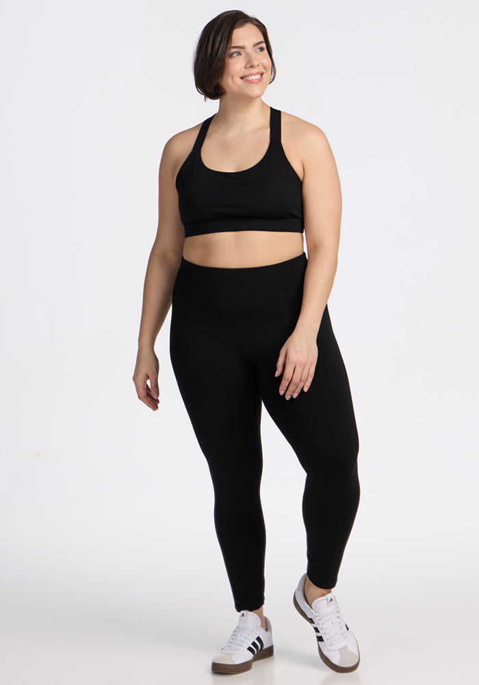 A woman wearing Woolx Angela Leggings - Black, a black sports bra, and white sneakers with black stripes stands smiling against a plain white background.