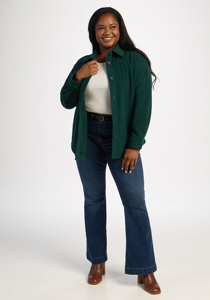 Woman standing and smiling, wearing the Woolx Jovie Shacket in Deep Pine over a white top, blue jeans, and brown boots.