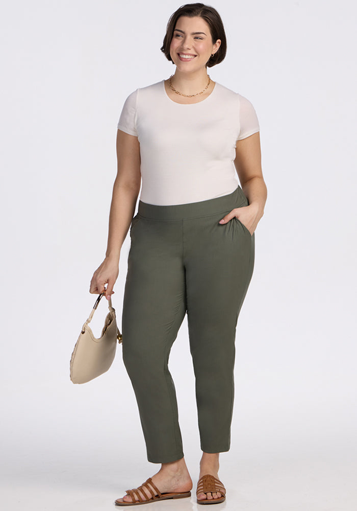A woman with short brown hair smiles, wearing a white short-sleeve top, Woolx Nicole Ankle Pants in Sage Olive, brown sandals, a necklace, and holding a beige handbag. The background is plain white.