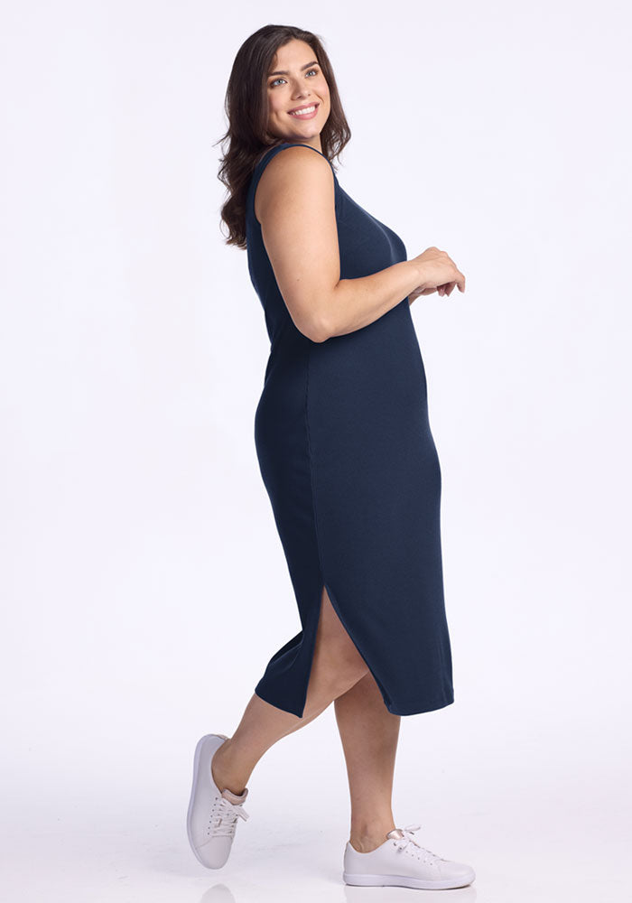 A woman with brown hair stands sideways, smiling in the Woolx Iris Ribbed Dress - Deep Navy. The sleeveless Merino wool dress features a side slit and pairs with white sneakers—an ideal travel-friendly choice against a plain white background.
