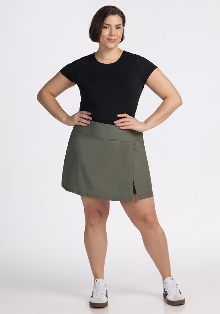 A woman with short brown hair stands facing forward against a white background, smiling slightly. She wears a black t-shirt, the Woolx Topanga Skort in Sage Olive with built-in shorts and pockets, and white sneakers, her left hand on her hip.