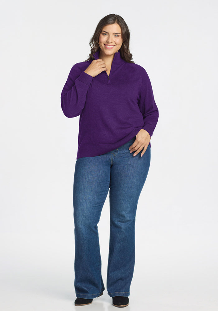 A woman smiles against a plain white background, wearing the Woolx Collins Half Zip Sweater in Plum Crush, blue jeans, and black shoes. One hand rests on her hip while the other touches her neck.