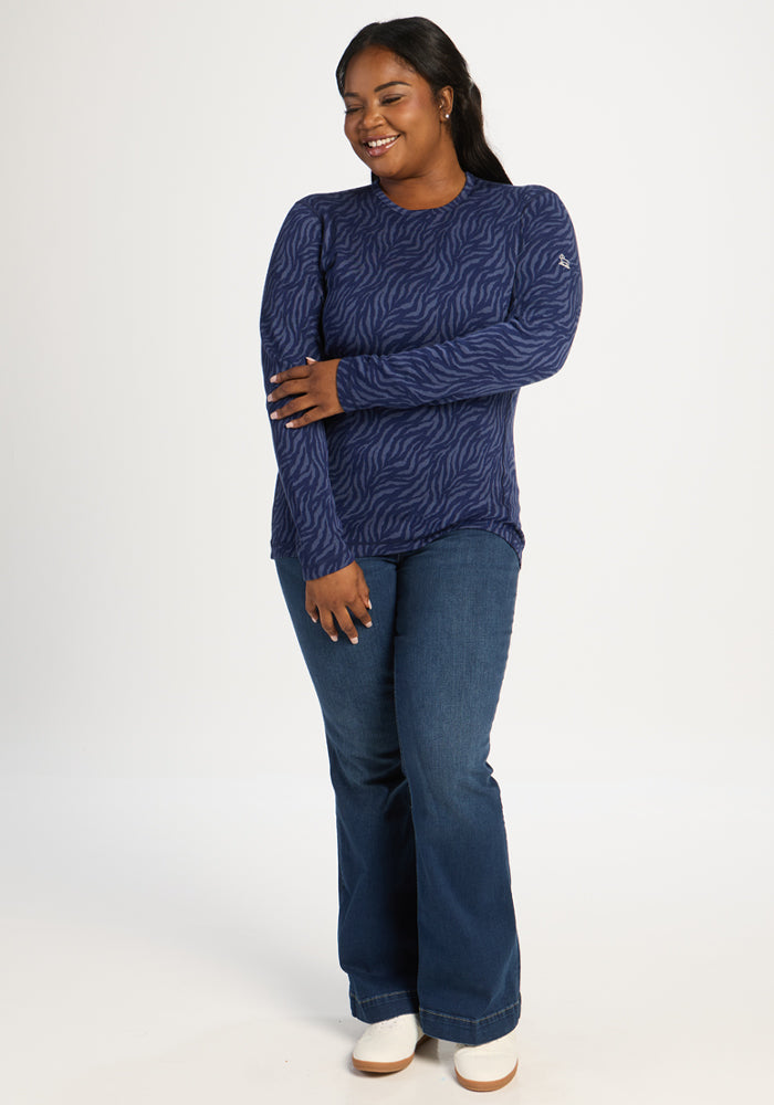 Woman wears Woolx Maddie Baselayer Top in Midnight Safari with blue jeans and white shoes, smiling against white backdrop.