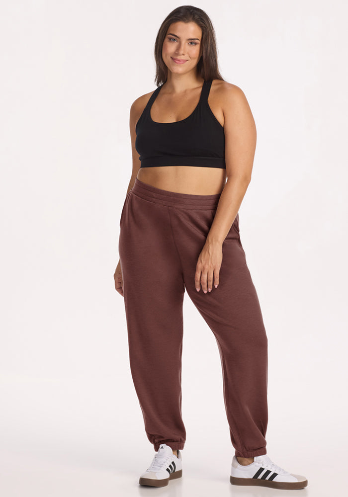 Woman in Parker Sweatpants - Canyon Clay by Woolx, black sports bra, and white sneakers, smiling against a plain background.