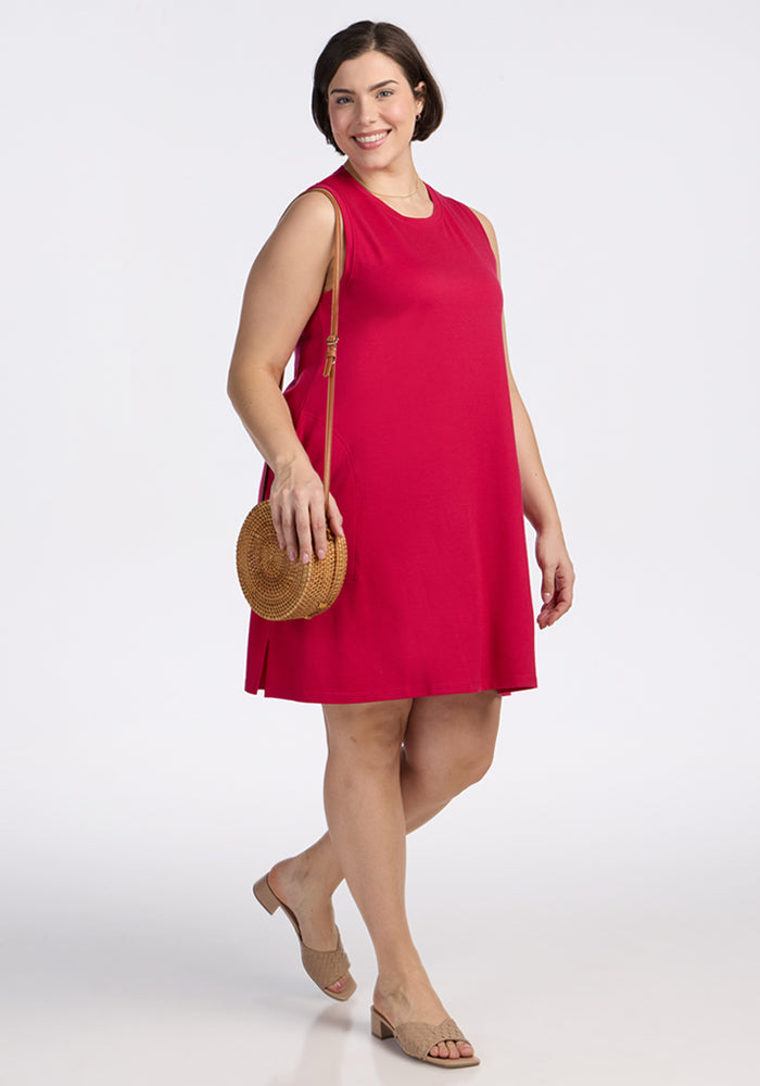 A woman in the Woolx Clara Dress in Poppy Spark, with beige sandals and a round woven crossbody bag, stands smiling against a plain white background.