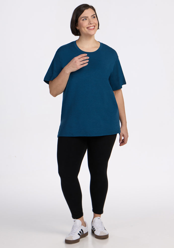 A woman with short brown hair wears the Woolx Audrey Tee in Majolica Blue, paired with black leggings and white sneakers with black stripes. She stands smiling, looking to the side against a plain white background.
