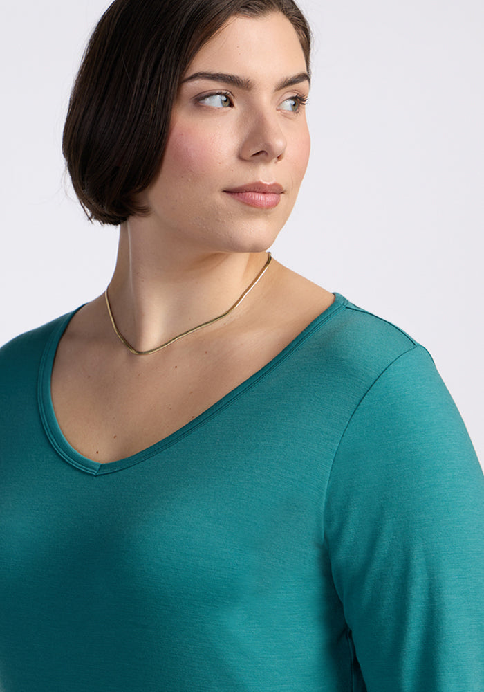 A person with short brown hair, wearing the Woolx Layla V Neck Top in Surfside and a gold necklace, looks to the side against a plain light background.