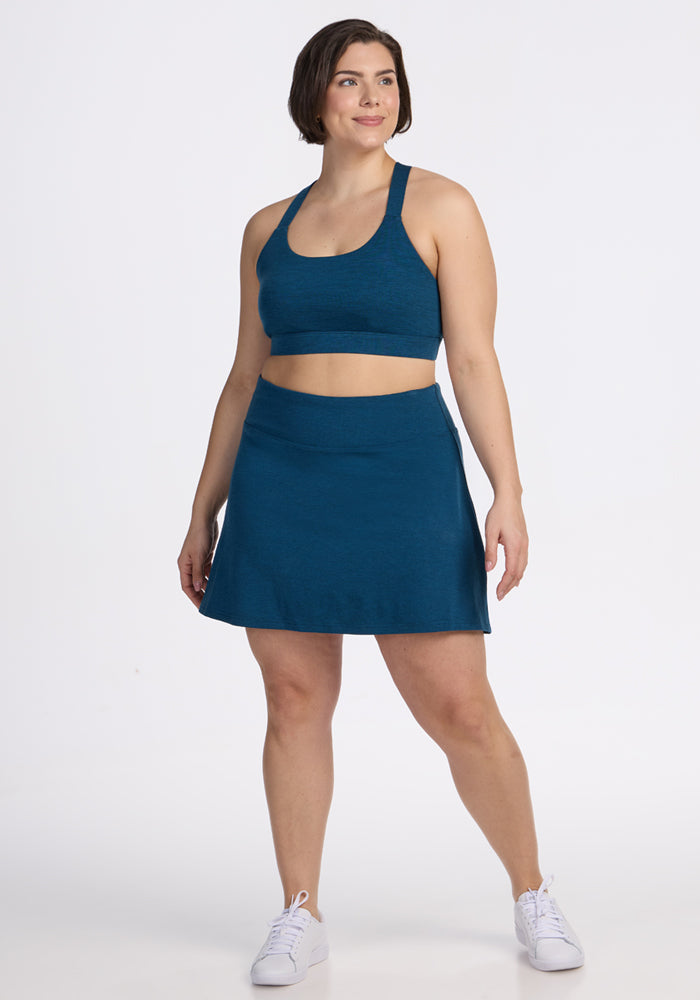 A woman with short brown hair smiles in a Majolica Blue Sienna Skort and matching sports bra by Woolx, paired with white sneakers, against a plain white background. 
