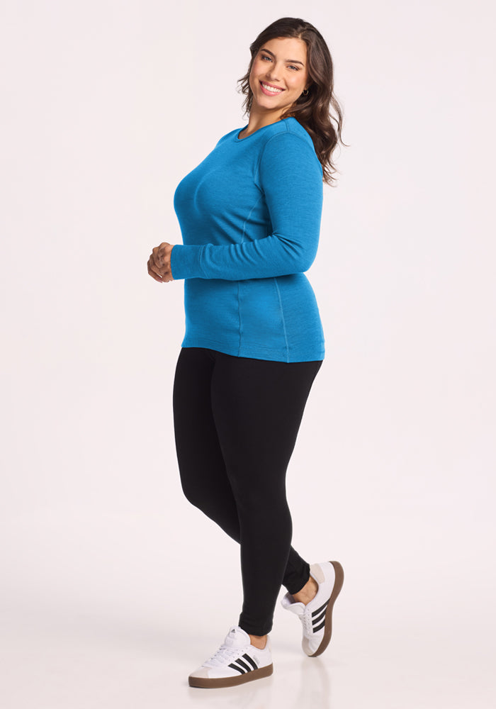 Woman wears the Woolx Hannah Long Sleeve Top in Blue Razz with black leggings and sneakers, smiling against white.