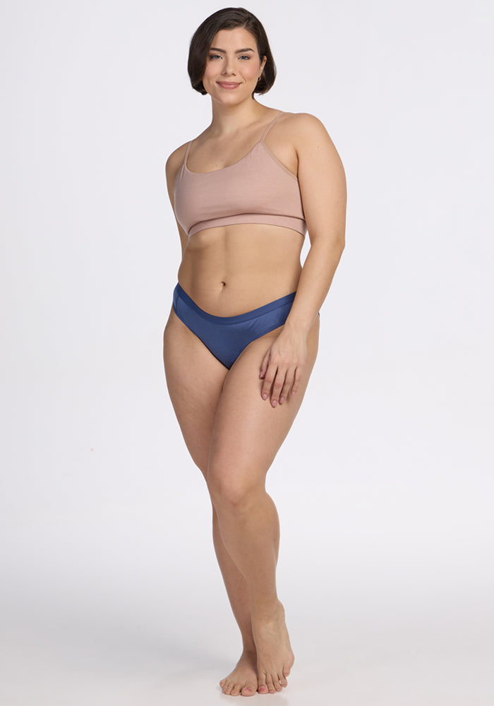 A woman with short brown hair stands barefoot against a plain white background, smiling with one arm relaxed by her side. She wears a light pink crop top and Woolx Roxie Bikini Underwear in the Skyline color.