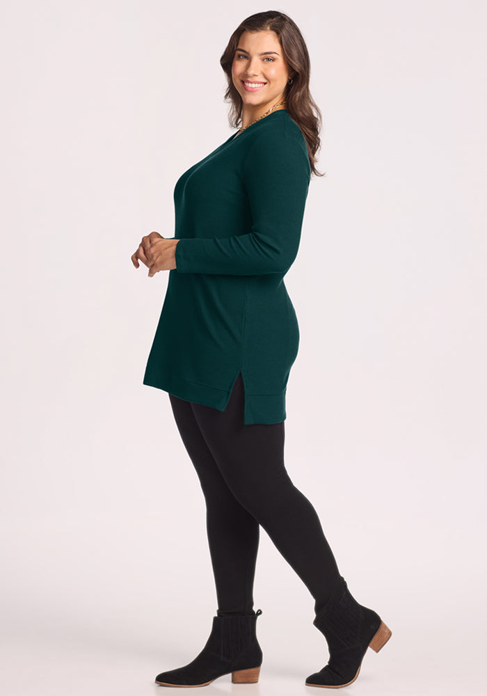 Woman in Woolx Evie Ribbed Tunic in Deep Pine, black leggings, and ankle boots stands smiling, facing sideways.