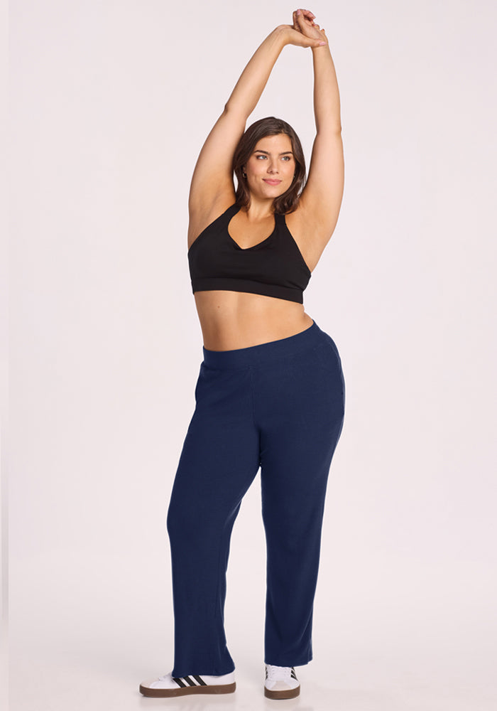 Woman in a black sports bra and Woolx Luca Ribbed Pants - Ocean Cavern stretches with arms overhead on a white background.