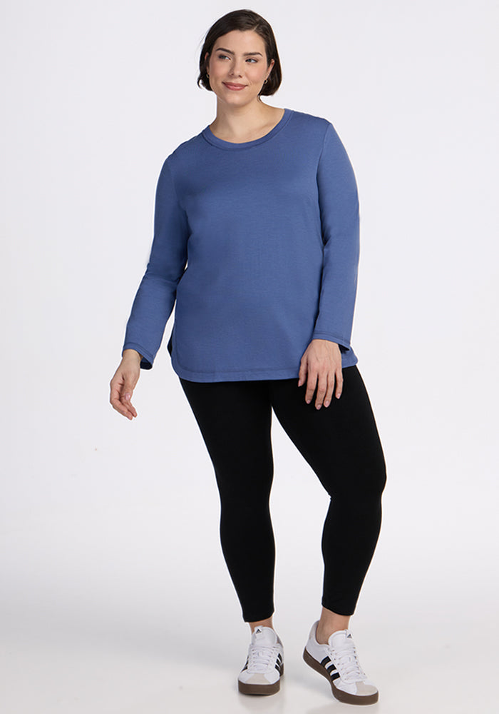 A woman with short brown hair wears the Woolx Hazel Tunic in Skyline, styled with black leggings and white sneakers featuring black stripes. She stands against a plain white background, smiling gently.
