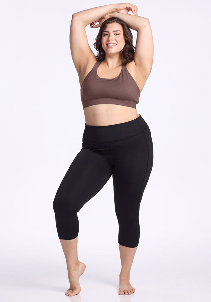 Woman in the Woolx Livi Bra - Peppercorn poses barefoot in black leggings, smiling with arms raised against a plain background.