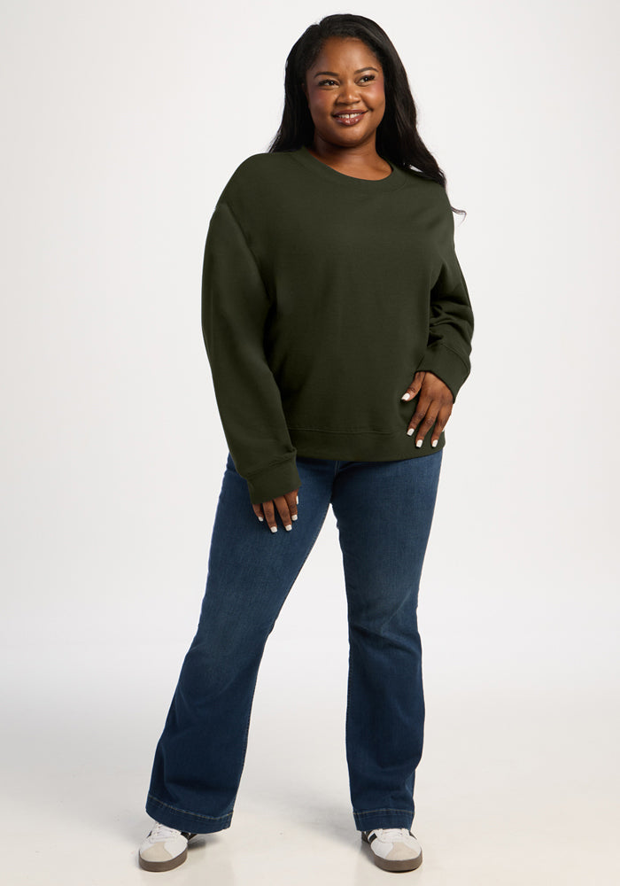 A woman wearing a dark green Molly Crewneck - Duffel Bag merino wool sweatshirt and blue jeans.