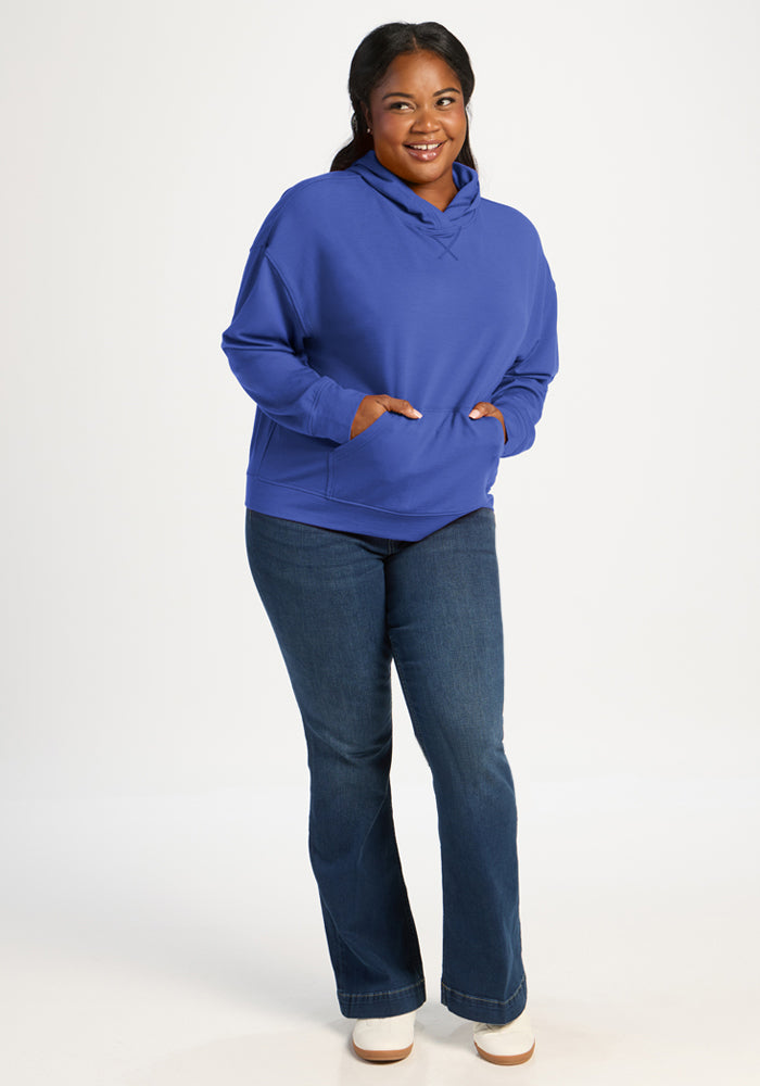 Woman wears Woolx Lizzy Hoodie in royal blue and jeans, smiling hands-in-pockets against a plain white background.