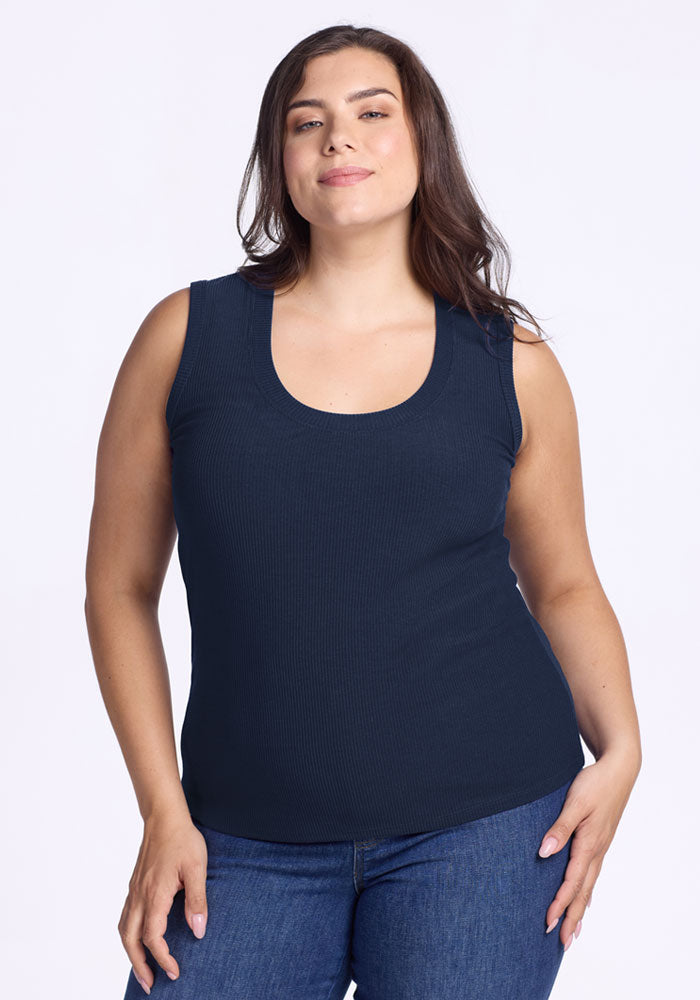 A woman with long brown hair wears the Woolx Sloane Ribbed Tank in Deep Navy and blue jeans, standing against a white background and smiling confidently with her hands relaxed by her sides. 