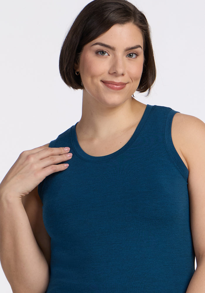 A woman with short brown hair gently smiles at the camera, wearing the Woolx Ava Tank Top in Majolica Blue, her hand raised near her shoulder against a plain light background.