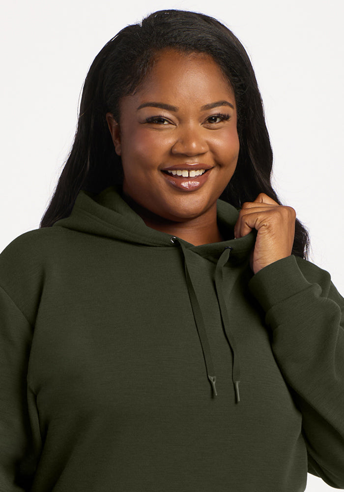 Smiling woman in a Woolx Avery Hoodie - Duffel Bag holds the drawstring, looking at the camera against a light background.