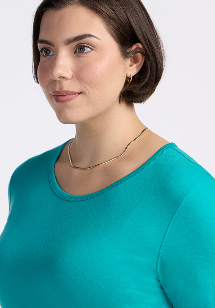 A woman with short brown hair wears the Woolx Remi Long Sleeve T-Shirt in Cool Jade, paired with a gold necklace and small hoop earrings. She gazes slightly to the side against a plain light background.