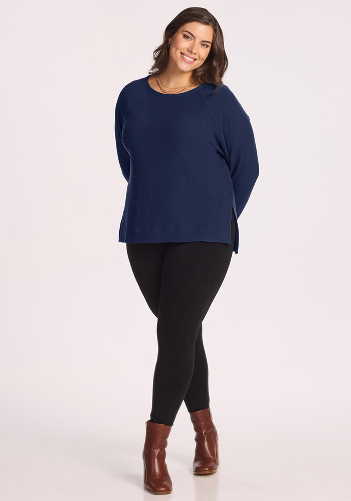 Woman smiles with hands behind her back, wearing the Woolx Ainsley Sweater in Ocean Cavern, black leggings, and boots.