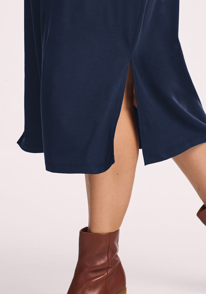 A person wearing the Woolx Cassie Dress in Deep Navy, a wrinkle-resistant travel dress with a side slit, paired with brown ankle boots, stands against a light background.