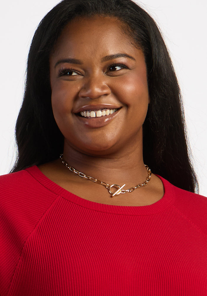 Smiling woman in Woolx Ainsley Sweater - Maraschino Cherry, with long dark hair and gold necklace against light background.