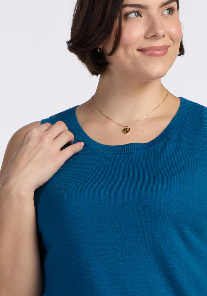 A person with short brown hair smiles while looking to the side, wearing a Woolx Chessy Tank Top in Moroccan Blue and a gold heart-shaped pendant necklace.