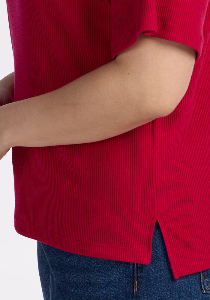A close-up shows a person in the Woolx Delia Ribbed Short Sleeve top in Poppy Spark, featuring merino wool and a side slit hem, paired with blue jeans. Only the arm, side, and waist are visible against a plain background.