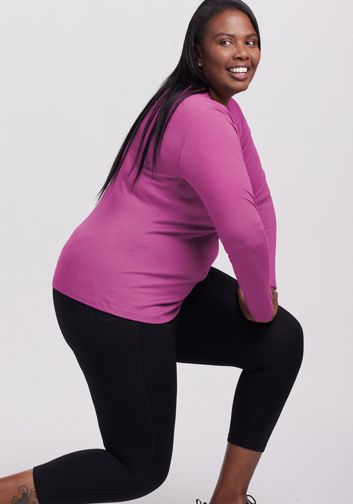 A smiling woman with long dark hair wears the Woolx Remi Long Sleeve T-Shirt in Purple Wine and black leggings, posing in a lunge against a plain light background.