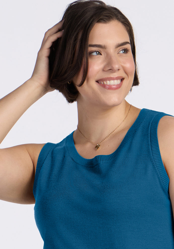 Smiling woman with short brown hair wears Woolx Chessy Tank Top in Blue Pine, touching her head with her left hand.