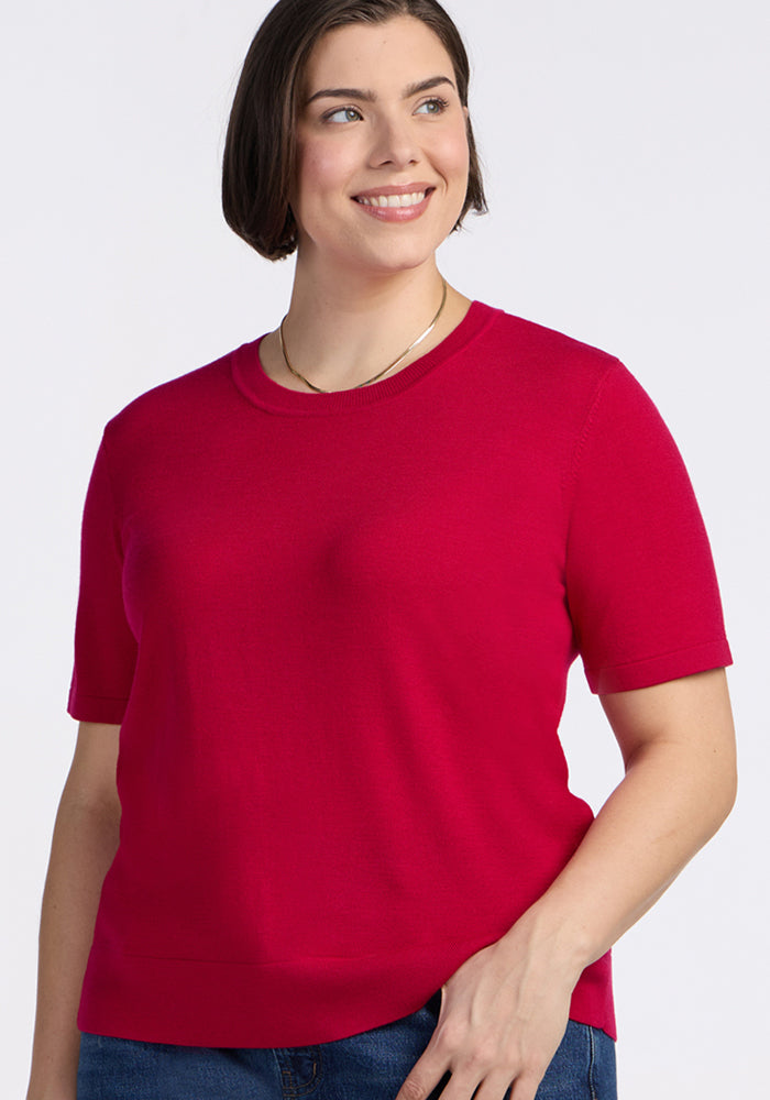 A woman with short brown hair smiles, wearing the Woolx Anna Crew Sweater in Poppy Spark and blue jeans, standing against a plain light background.