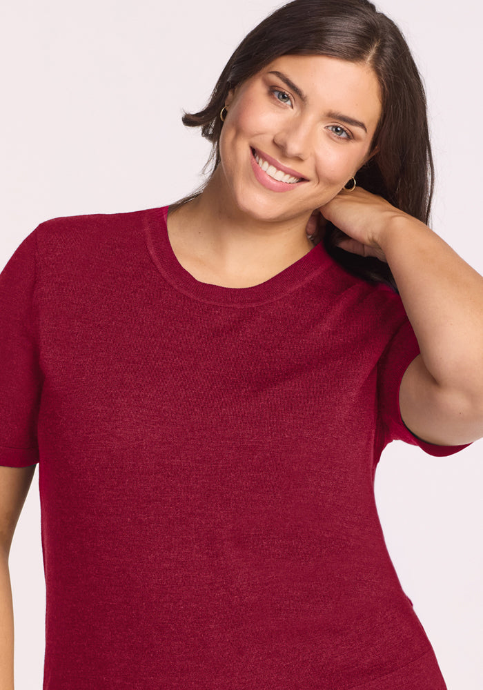 Woman with brown hair smiles, touching her neck in a Woolx Anna Crew Sweater - Beet Red against a light background.
