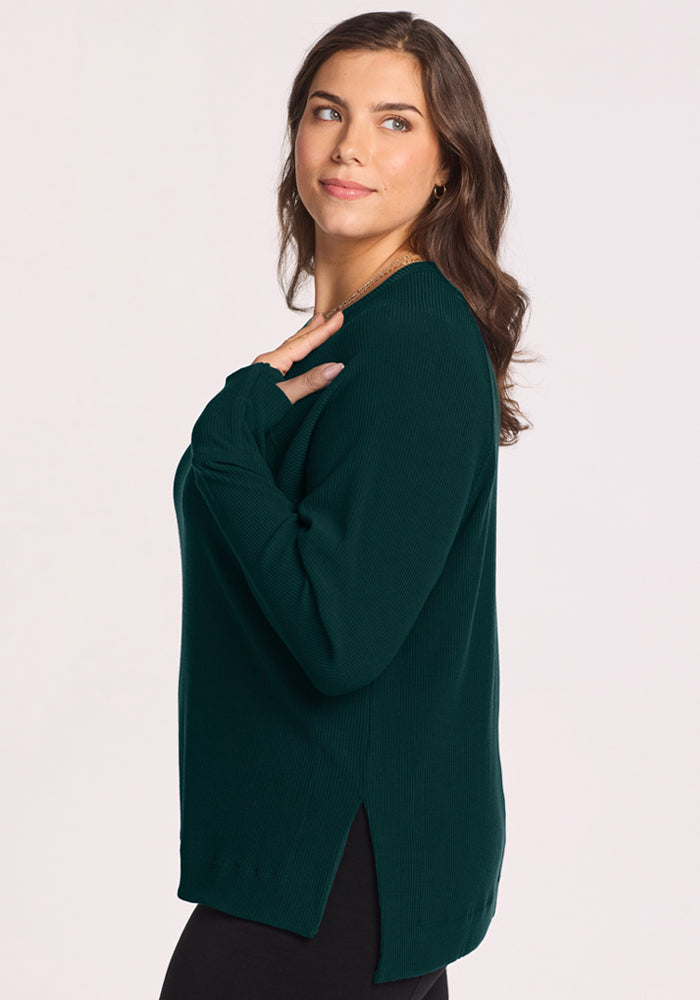 A woman in the Woolx Ainsley Sweater in Deep Pine stands sideways, glancing over her shoulder with a soft smile.