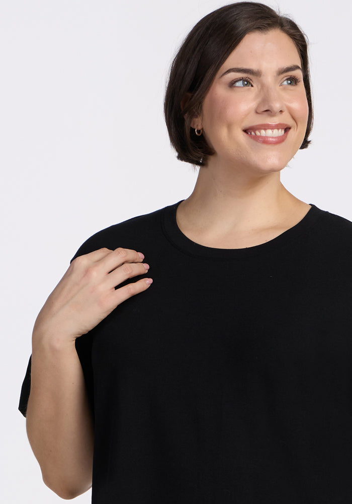 A woman with short brown hair wears the Woolx Audrey Tee in black, smiling and looking to the side against a plain light background, her left hand raised near her shoulder.