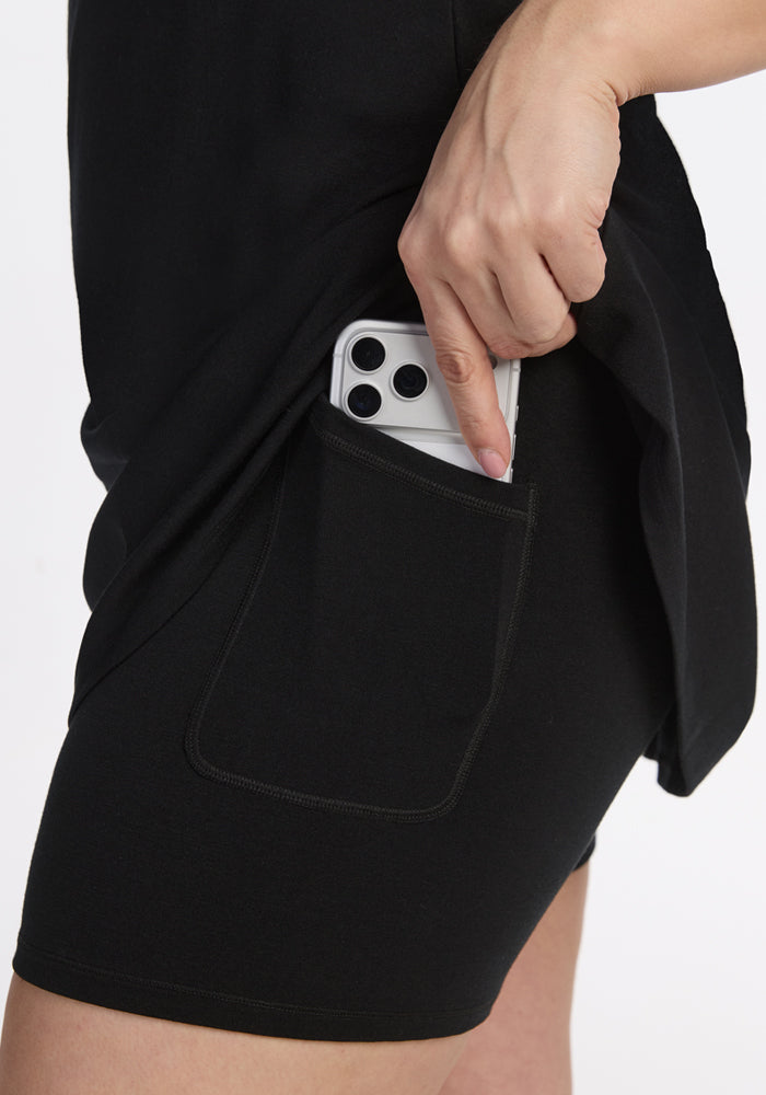 A person wearing the Woolx Adrienne Active Dress in black, which features built-in shorts, slips a triple-camera smartphone into the side pocket of the shorts.