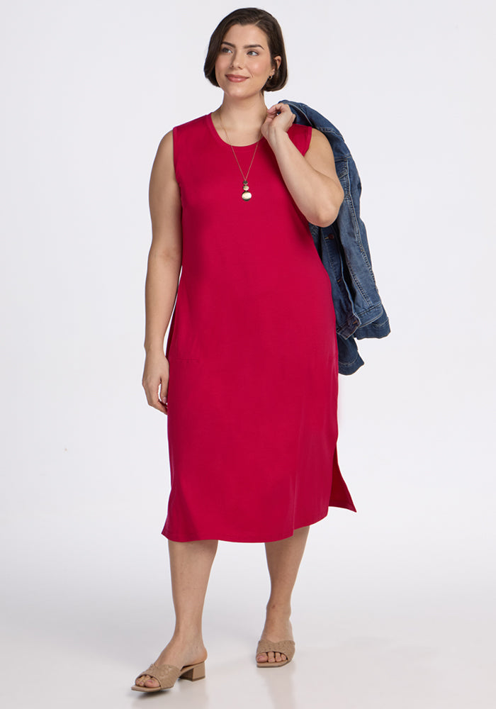 A woman smiles against a white background, wearing the Woolx Cassie Dress in Poppy Spark with beige sandals and a necklace, holding a denim jacket over her shoulder.