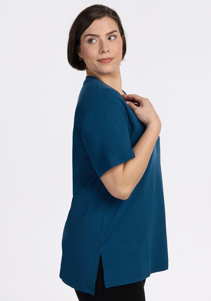 A woman with short dark hair models the Woolx Audrey Tee in Majolica Blue, paired with black pants, standing sideways and looking over her shoulder against a plain white background.