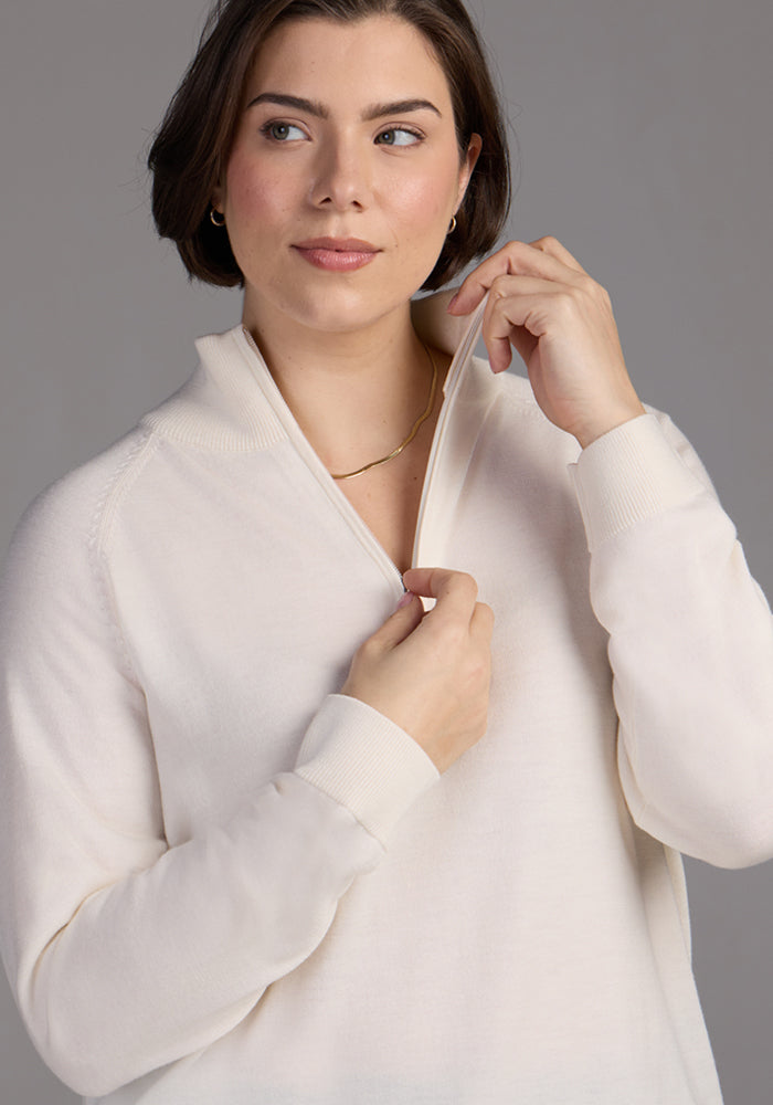 A woman with short brown hair wears the Woolx Collins Half Zip Sweater in Dewdrop Pearl and a thin gold necklace. She is pulling up the zipper, looking slightly to the side with a soft, neutral expression against a plain background.