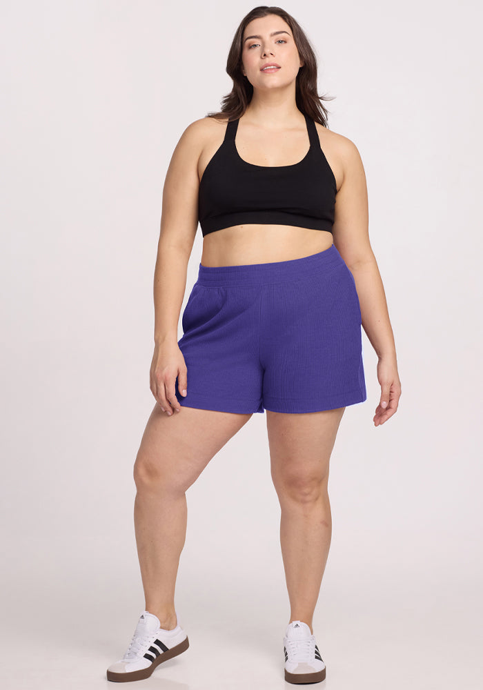 Woman in a black sports bra, Woolx Maisey Ribbed Shorts - Berry Indigo, and sneakers against a plain white background. 