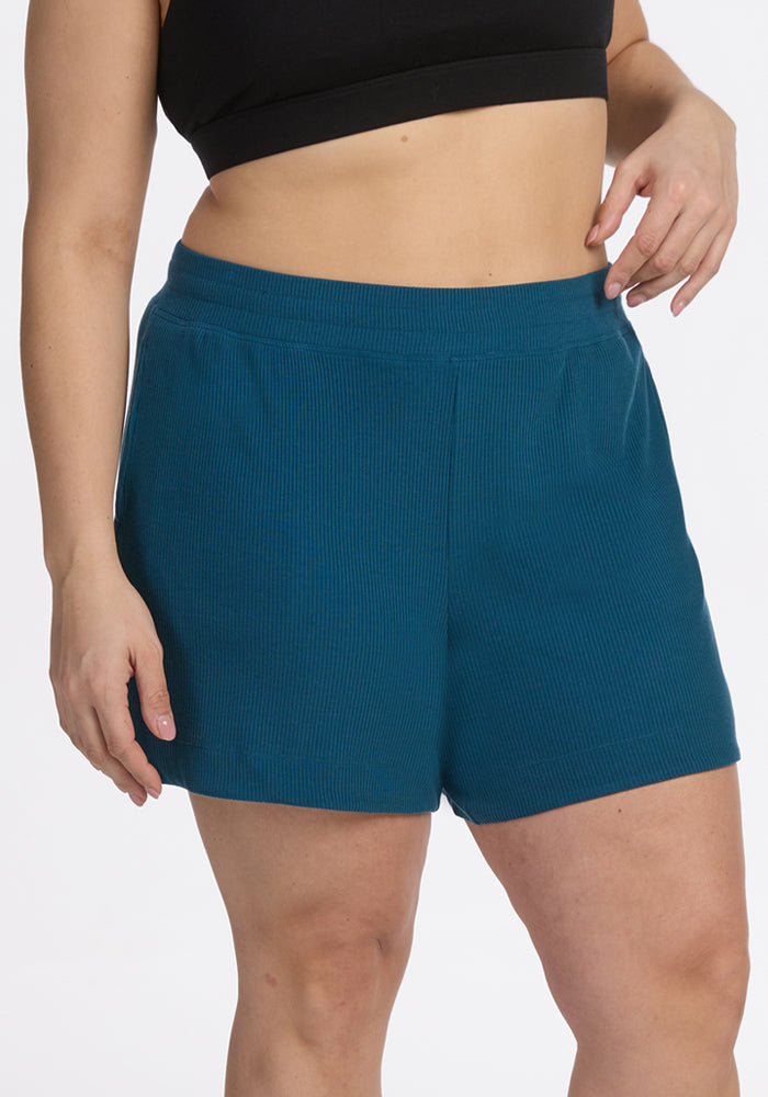 A person models Woolx Maisey Ribbed Shorts in Moroccan Blue and a black crop top against a plain white background, with one hand at their side and the other resting on their hip.