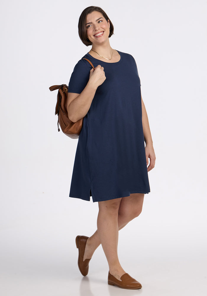 A smiling woman wears the Woolx Georgie Dress in Deep Navy, brown loafers, and carries a brown backpack while standing against a plain white background.