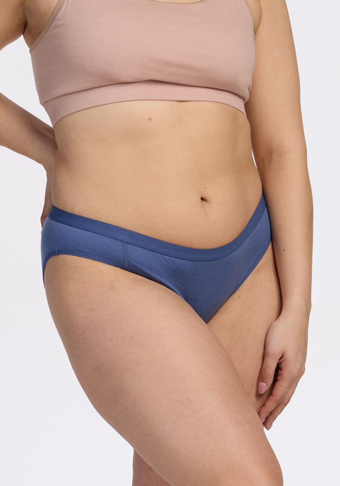 A person wears the Woolx Roxie Bikini Underwear in Skyline and a nude bra, standing against a plain white background with one arm at their side and the other hand on their thigh.