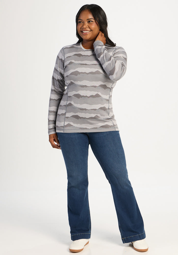 Woman in Woolx Maddie Baselayer Top - High Peaks and blue jeans smiles, one hand touching her hair.