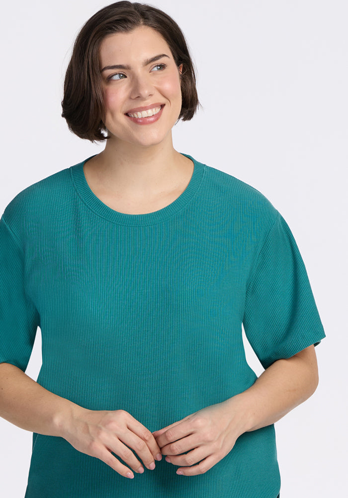 A woman with short brown hair smiles slightly to the side, wearing the Woolx Delia Ribbed Short Sleeve in Surfside. The relaxed fit merino wool top stands out against a plain light background.