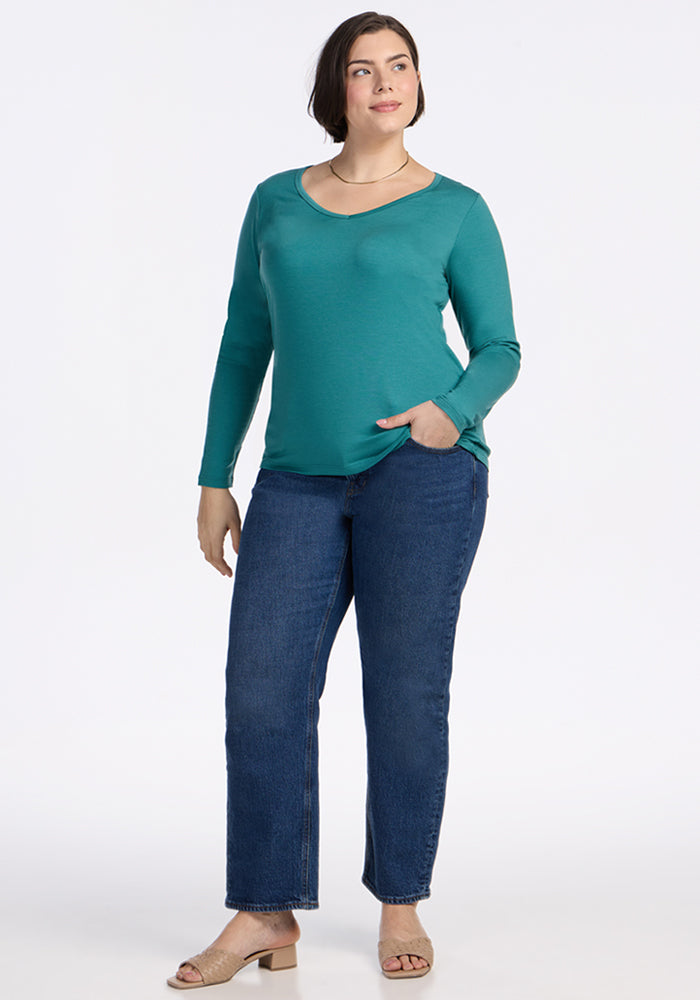 A woman with short dark hair smiles against a white background, wearing Woolx's Layla V Neck Top in Surfside, blue jeans, and beige heeled sandals. The odor-resistant merino wool top keeps her fresh all day with one hand in her pocket.