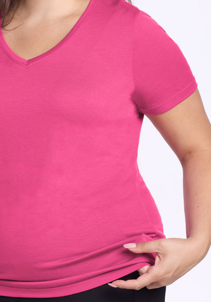 A person is shown from the torso up, wearing the Woolx Junie V Neck in Raspberry Fizz with black pants, holding the side of the shirt with their left hand. Only part of their right arm and upper body are visible.